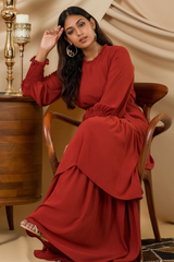 A woman wearing a rust-colored, long-sleeved shirt with a round neckline and a matching skirt with a midi-length, also in a rust color. They are standing in front of a beige fabric backdrop.